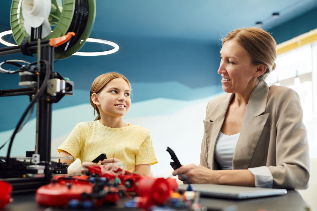 Courses Low angle of smiling woman and little girl sitting at table and working on engineering project together while looking at each other in light workspace